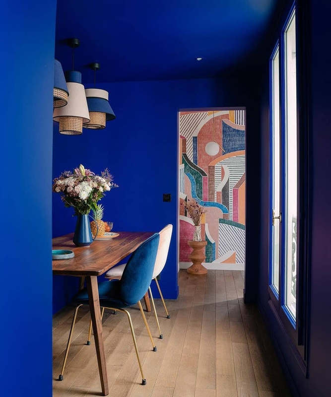 Parede da sala de jantar na cor Sombra Azul Suvinil e mesa com decorações de madeira. Ao fundo, painel colorido abstrato.