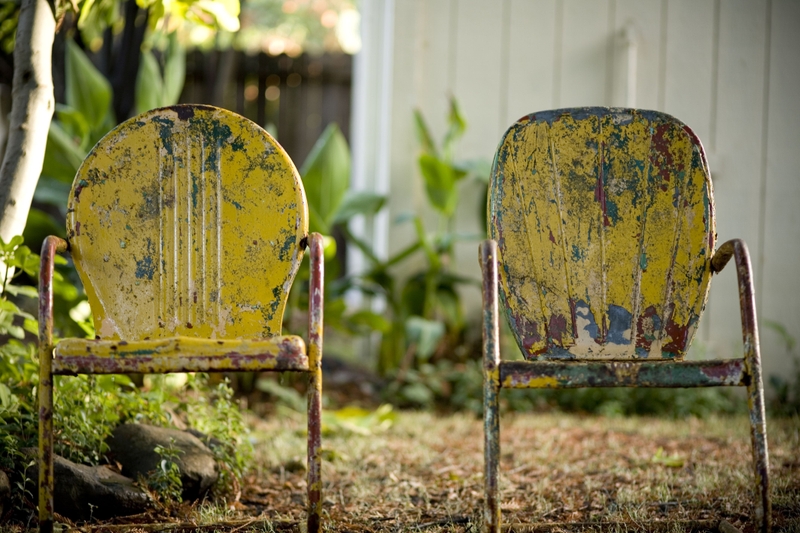 A foto mostra duas cadeiras amarelas com ferrugem e desgaste azul no jardim. Uma cadeira está à esquerda da imagem e a outra, à direita. Ao fundo e desfocadas, há árvores, plantas e a fachada de uma casa com ripas de madeira branca.