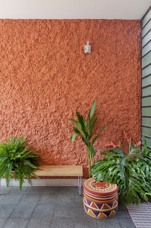  A foto mostra a parede lateral da área externa da entrada da casa. À esquerda, banco de madeira com plantas em cima e à direita, plantas sobre o piso de azulejo cinza, puff estampado colorido, tapete listrado e janela de vidro jateado com esquadria preta. Ao fundo, parede texturizada pintada com a cor Terra Roxa.