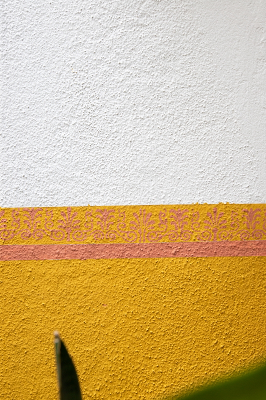 A foto mostra uma parede externa texturizada. A metade de cima da parede foi pintada de branco, já a metade debaixo recebeu a cor Quentão com uma linha e arabescos desenhados com a cor Terra Roxa.