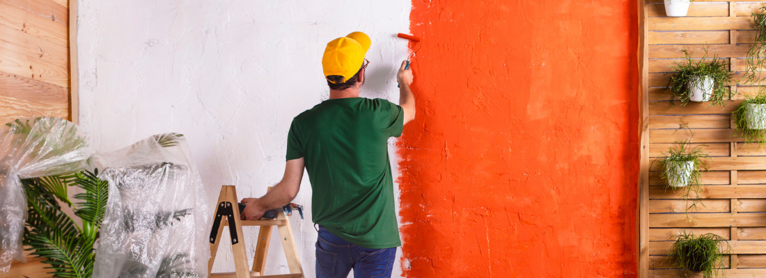 Homem pintando, com um rolo de pintar, uma parede da cor laranja.
