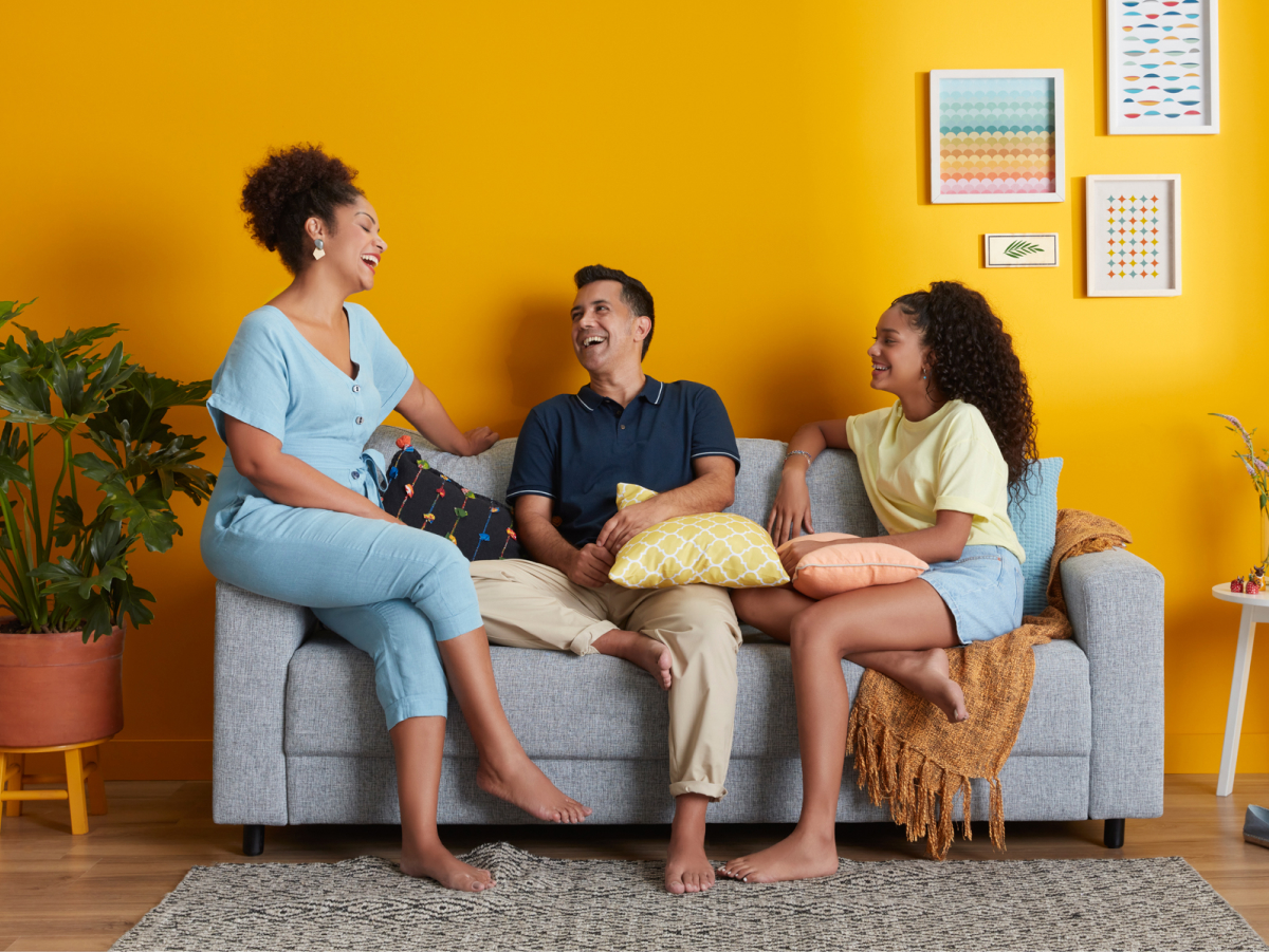Família sentada no sofá e sorrindo. Atrás deles tem uma parede laranja e um quadro colorido.
