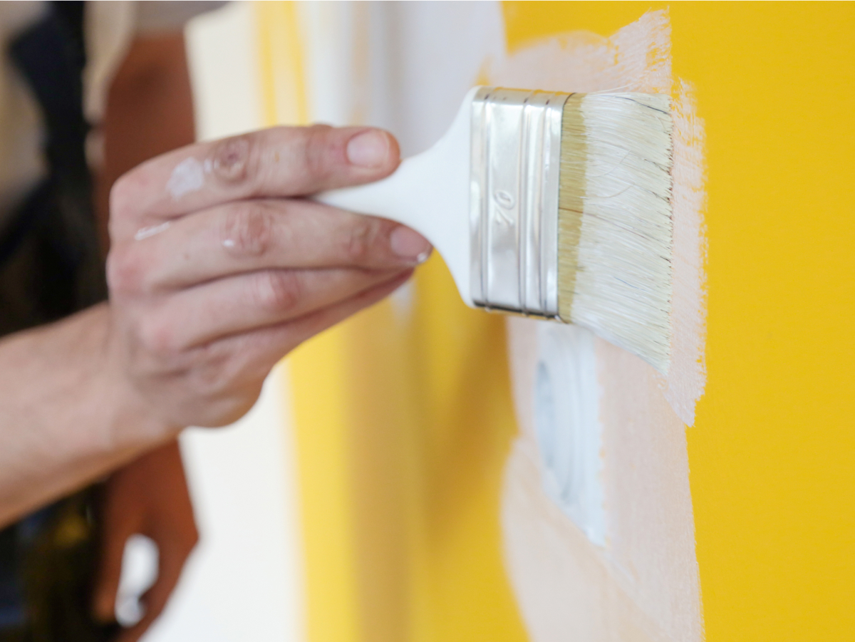Parede amarela sendo pintada de branca. Apenas a mão do pintor, segurando o pincel, aparece na foto.
