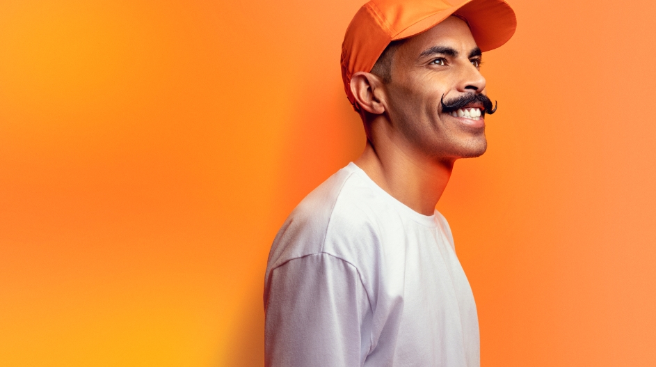 Homem, usando um boné laranja, sorrindo e olhando para a sua frente.