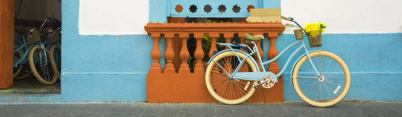 Bicicleta azul com rodas brancas apoiada em uma pareda colorida em uma parede azul de tonalidade mais clara, com detalhes em laranja.