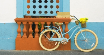 Bicicleta azul com rodas brancas apoiada em uma pareda colorida em uma parede azul de tonalidade mais clara, com detalhes em laranja.