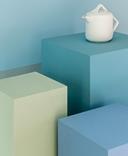 Um bule branco, de design moderno, está posicionado em cima de um de três pilares, cada qual em um tom diferente de azul, representando a versatilidade desta matiz.